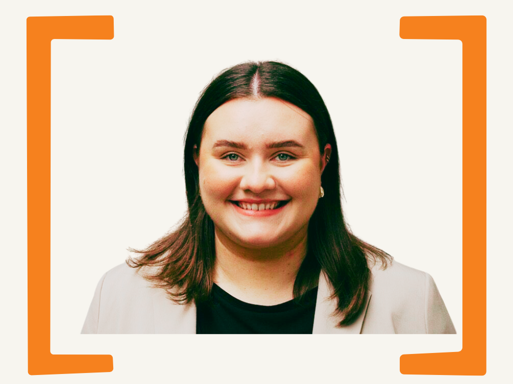 A smiling woman with shoulder-length dark hair wearing a beige blazer and black top against a white background with orange border accents.