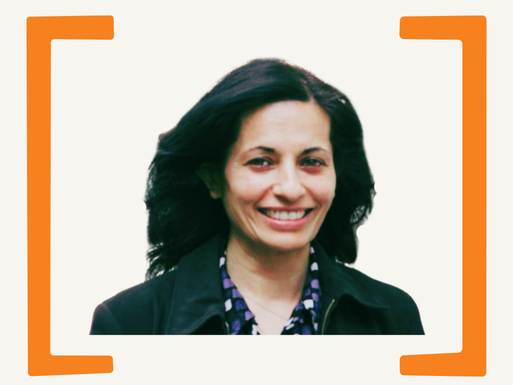 A woman with dark hair smiling, wearing a black jacket and a patterned blouse, on a light background with orange border corners.