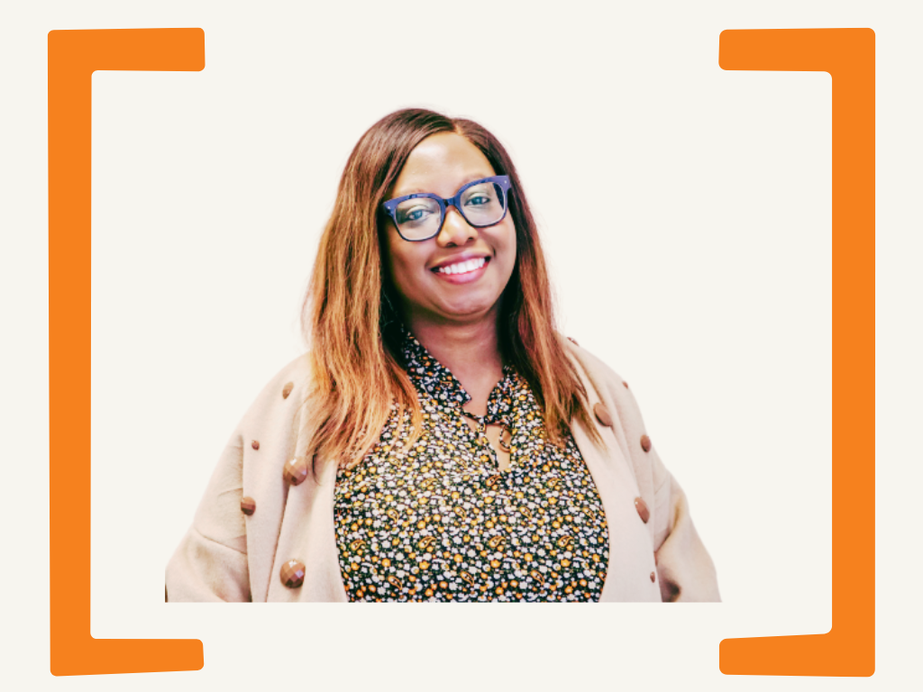 A woman with shoulder-length brown hair, glasses, and a smile, wearing a patterned shirt and a beige jacket, posing against a white background with orange border accents.